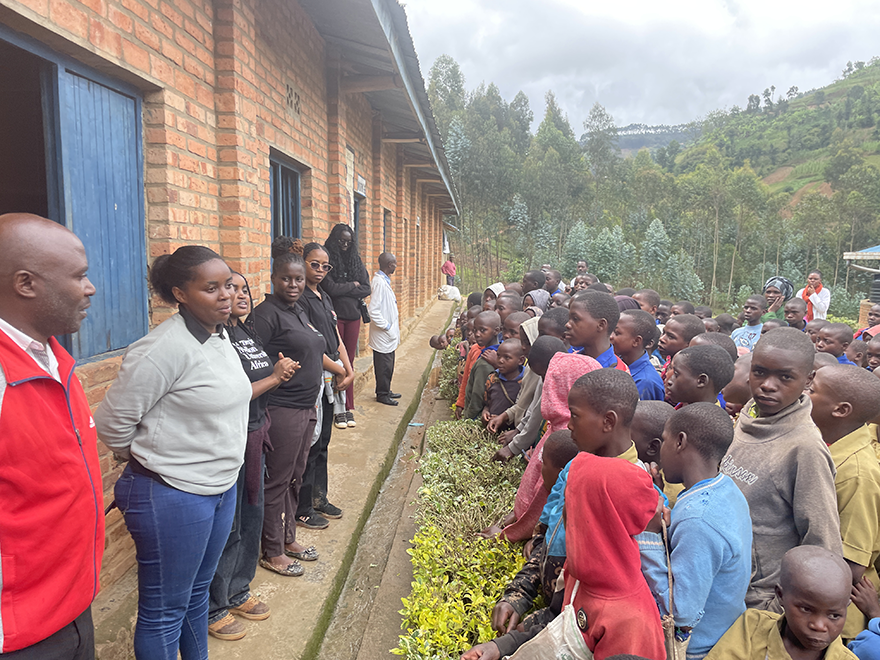 A group of adults engages with children outside a classroom building in a rural setting. The scene highlights community interaction and educational outreach.