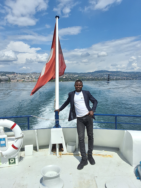 A man posing on a boat with a waving flag and scenic background of water and hills.