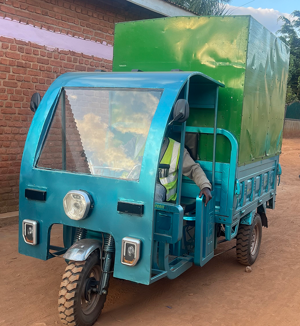 A blue three-wheeled cargo truck with a green covered box on the back, parked on a dirt road beside a brick building.