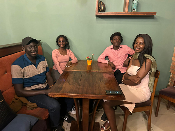Four friends sit around a wooden table in a cozy café booth, smiling at the camera, with a small potted plant centered on the table.
