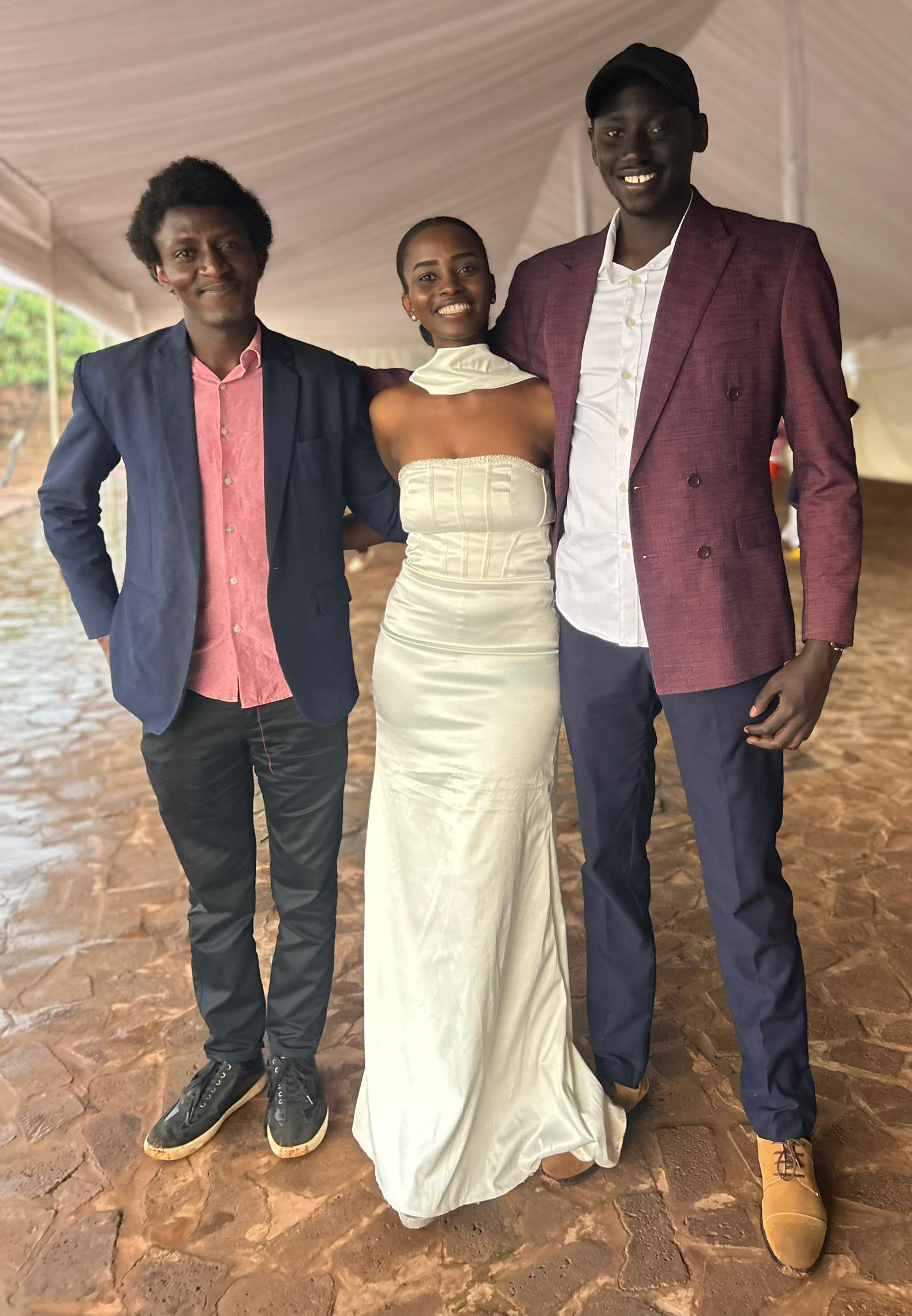 Three people pose smiling under a white event tent; the woman in the middle wears a strapless white dress, with two men in blazers standing on either side.