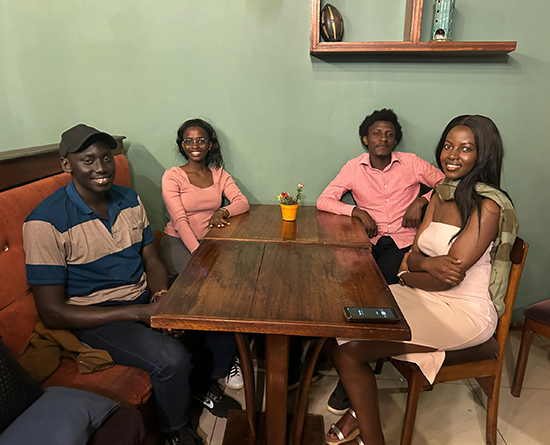 Four friends sit around a wooden table in a cozy café booth, smiling at the camera, with a small potted plant centered on the table.